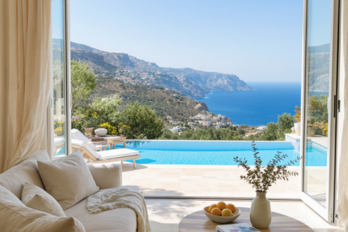 Vue panoramique depuis une villa de luxe en Crète - salon ouvert sur piscine à débordement et mer Égée, intérieur design avec canapé beige et table basse en bois naturel, ambiance lumineuse et apaisante